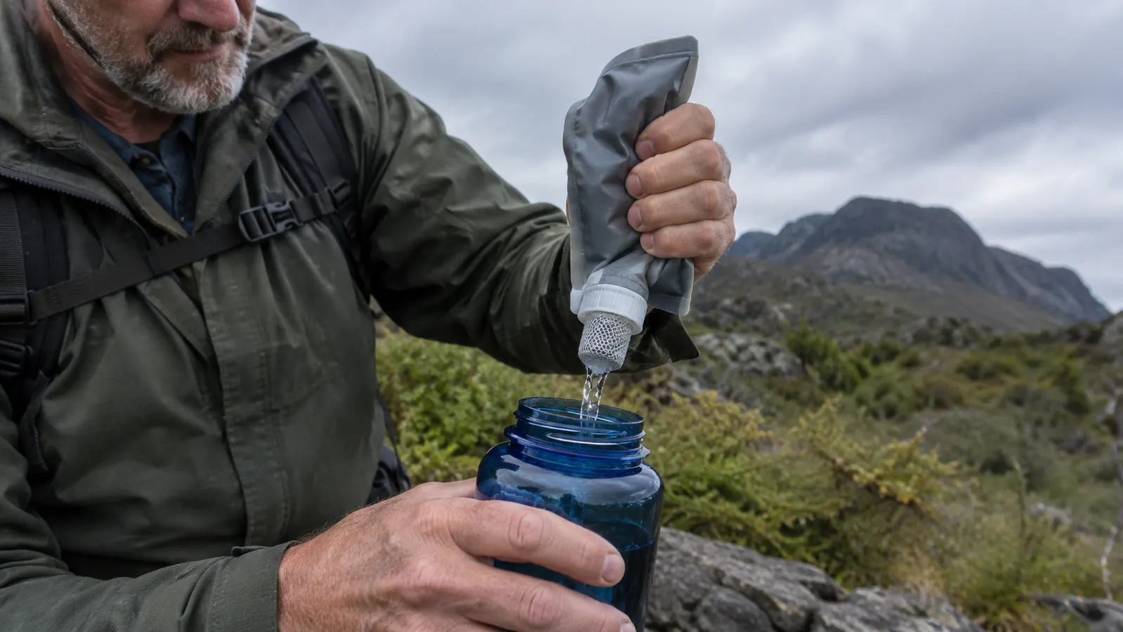 A hand squeezing a small filter pouch over a wide-mouth water bottle on a trail